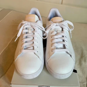 Vita sneakers från Adidas - Säljer ett par nya snygga, supersköna vita Adidas Advantage sneakers.  Mycket skön sula - cloudfoam.  Skorna har en stilren design med snörning och en diskret grön detalj på hälen. Säljes pga felköp. Stl 38