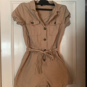 Beige jumpsuit med knappar - Säljer en snygg beige jumpsuit med korta ärmar och knappar framtill. Den har en knytning i midjan för en justerbar passform och två bröstfickor med knappar. Perfekt för en avslappnad stil.