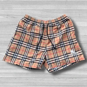 Burberry shorts - Säljer ett par klassiska rutiga badshorts från Burberry i beige, svart och röd. Shortsen har elastisk midja och en praktisk bakficka. Perfekta för sommarens stranddagar! Strolek: XL men passar även M