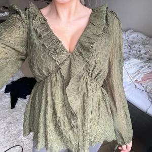Olivgrön blus med volangdetaljer - Säljer en olivgrön blus med v-ringning och volangdetaljer. Blusen har långa ärmar och ett omlottmönster som ger en elegant look. Perfekt för både vardag och speciella tillfällen. Passar strl S/36