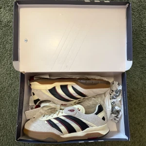 Adidas x SNS - Snygga Adidas sneakers i beige med svarta och röda ränder. Skorna har en klassisk design med snörning och en bekväm gummisula. Perfekta för en stilren och sportig look.