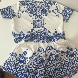 Vit och blå mönstrad topp och shorts - Säljer en snygg vit och blå mönstrad topp med matchande shorts. Toppen har korta ärmar och ett detaljerat blommönster i blått. Perfekt för sommardagar!