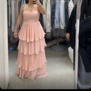 Rosa långklänning med volanger - Elegant rosa långklänning med flera lager av volanger och smala axelband. Klänningen har en romantisk känsla med sin mjuka färg och flytande design. Perfekt för speciella tillfällen. Har använd en gång 