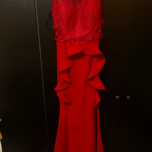 Röd långklänning med spetsdetaljer - Elegant röd långklänning med spetsdetaljer och volanger. Klänningen har en v-ringad design och är ärmlös, vilket ger en stilren look. Perfekt för festliga tillfällen.