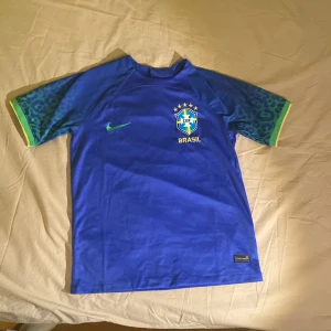 Blå Brasilien fotbollströja från Nike - Säljer en blå Brasilien fotbollströja från Nike med korta ärmar och gröna detaljer. Tröjan har Brasiliens emblem på bröstet och en grön Nike-logga. Perfekt för fotbollsfans! 🇧🇷