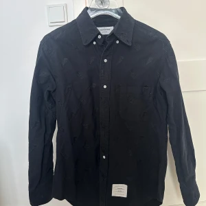 Svart skjorta från Thom Browne - Snygg svart skjorta från Thom Browne med subtila broderade detaljer. Skjortan har en klassisk krage och knappar framtill. Perfekt för en stilren look.