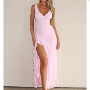 Rosa balklänning från white fox - Elegant rosa långklänning med v-ringning och volangdetaljer längs slitsen. Klänningen är ärmlös och har en smickrande passform som framhäver figuren. Aldrig använd! 