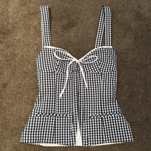 Svartvit rutig korsettopp - Säljer en snygg svartvit rutig korsettopp med knytband framtill. Toppen har breda axelband och en söt rosett vid bysten. Ny skick och aldrig använd. Perfekt för sommaren 💕