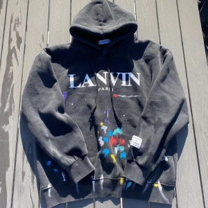 Svart hoodie från Lanvin - Säljer en svart hoodie från Lanvin med färgglada stänk och texten 'LANVIN PARIS' på framsidan. Baksidan har en diskret text 'DÉPT de la GALERIE'. Hoodien har en etikett med 'ART ON DISPLAY Courtesy of JOSUÉ THOMAS'. Perfekt för dig som gillar unik design!