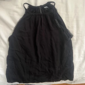 Svart ärmlös linnetopp med spetsdetaljer - Svart ärmlös linnetopp med tunna axelband och dekorativa spetsdetaljer längs sidorna. Toppen är från hm. Lätt rynkad struktur och en liten öppning bak vid halsen. 