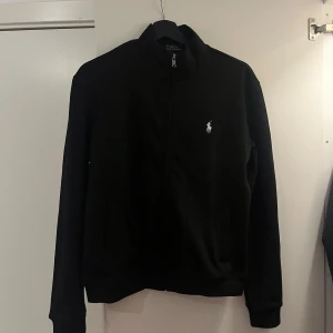 Svart tröja från Polo Ralph Lauren M - Svart långärmad tröja från Polo Ralph Lauren i custom fit. Klassisk design med dragkedja framtill och vit broderad logga på bröstet. Perfekt för dig som gillar stilrena och tidlösa plagg. Tillverkad i mjuk bomull för skön känsla. Prutning kan ske vid seriös köpare. Storlek M men passar oxå S. Beror egentligen på kroppsuppbyggnaden. 