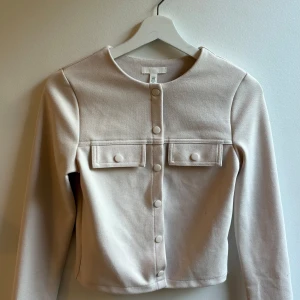 Beige overshirt från H&M XS - Snygg beige overshirt från H&M i storlek XS. Jackan har raka linjer, rund halsringning, tryckknappar framtill och två dekorativa bröstfickor. Perfekt att slänga över en t-shirt för en clean och stilren look.