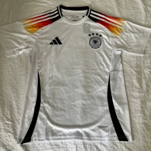 Tysklands fotbollströja Adidas - Vit Tysklands fotbollströja från Adidas med svarta detaljer och meshpaneler på sidorna. Axlarna har ränder i svart, rött och gult. Broderad landslagslogga och Adidas-märke på bröstet. Tillverkad i lätt och ventilerande material.