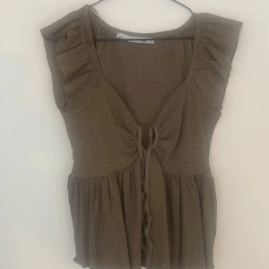 Brun blus med volang och knyt - Supersöt brun topp från lager 157  med volangdetaljer vid axlarna och knyt framtill. Toppen  har v-ringning och är ärmlös, med lätt rynkat tyg som ger en luftig känsla. Perfekt för dig som gillar en   trendig stil.