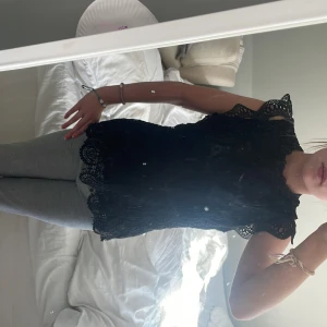 Svart spetsblus med volangärm - Supersnygg svart blus i spets med detaljerat paisleymönster och volang vid ärmslut. Blusen är ärmlös med hög hals och knäppning bak i nacken. Perfekt för dig som gillar romantisk och trendig stil.