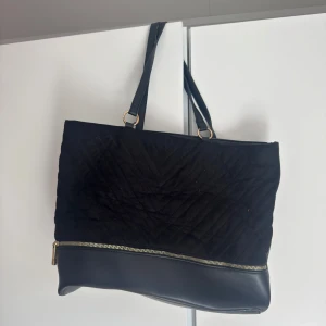 Svart axelväska - Stilren svart axelväska med quiltad ovandel och slät botten i skinnimitation. Väskan har flera fack inuti och detaljer i guld, samt en synlig dragkedja framtill. Perfekt för dig som vill ha plats för allt du behöver under dagen. Dock är väskan lite sliten på axelremmen (se bild två). Pris kan diskuteras.