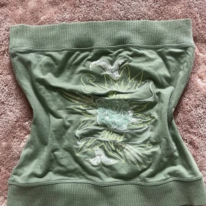 Grön tubtopp från MNG Jeans med paljetter - Cool grön tubtopp från MNG Jeans med broderad text 'Free Bird', blommigt motiv och glittriga paljettdetaljer framtill. Tjocka ribbade kanter upptill och nedtill ger en snygg passform. Perfekt för dig som vill sticka ut med en unik och trendig look.