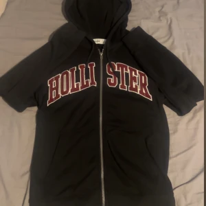 Svart Hollister hoodie med dragkedja - Svart hoodie från Hollister med stor röd och vit logga över bröstet. Tröjan har huva, korta ärmar och hel dragkedja framtill. Perfekt för en chill och sportig look. Fickor framtill för extra bekvämlighet.