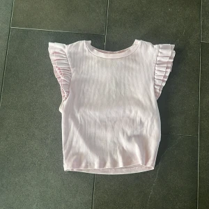 Ljusrosa volangtopp från Zara - Supersöt ljusrosa topp från Zara i ribbad bomull. Toppen är ärmlös med breda volangdetaljer på axlarna som ger en extra touch. Perfekt för dig som gillar en feminin och trendig stil. Enkel att matcha med både jeans och kjol.