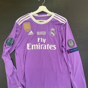 Real Madrid Ronaldo CL-tröja - Säljer en ikonisk långärmad Real Madrid-tröja från Champions League-finalen 2017 i Cardiff. Lila med vita detaljer, Adidas-logga, klubbmärke, FIFA-märke och tryck för Ronaldo 7 på ryggen. Materialet är lätt och andas, perfekt för fotbollsfans.