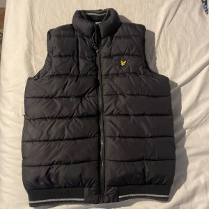 Svart dunväst från Lyle & Scott Junior - Snygg svart dunväst från Lyle & Scott Junior med gul logga på bröstet. Västen är quiltad med horisontella sömmar, har hög krage och dragkedja framtill. Ribbad kant nertill med vit rand för extra stil. Perfekt för lager-på-lager under kyliga dagar.