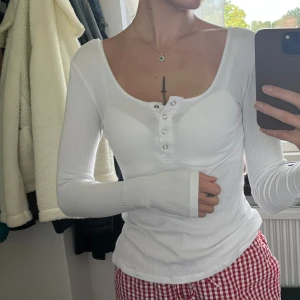 Vit ribbad långärmad topp Pieces - Säljer en vit ribbad långärmad topp från Pieces med snygg v-ringning och knappdetaljer framtill. Tröjan är figurnära och har stretchigt material som sitter skönt på kroppen. 