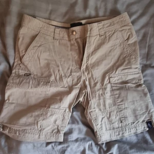 Beige cargoshorts från Five Seasons - Snygga beige cargoshorts från Five Seasons med flera praktiska fickor på sidorna och bak. Klassisk passform med bälteshällor och knappstängning. Perfekta för sommardagar och äventyr utomhus. Lätta och sköna i bomullsmaterial.