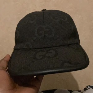 Svart keps från Gucci med GG-mönster - Snygg svart keps från Gucci med klassiskt GG-mönster ton-i-ton över hela kepsen. Bred skärm med svart kant och justerbar rem baktill. Tillverkad i ett slitstarkt tygmaterial som ger en exklusiv känsla.