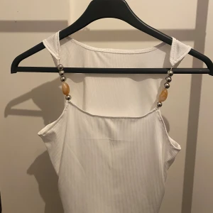 Vit ribbad linnetopp från SHEIN - Supersnygg vit ribbad linnetopp från SHEIN i storlek S. Toppen har smala axelband med coola pärldetaljer i silver och beige, vilket ger en unik touch. Perfekt att styla med jeans eller kjol för en trendig look. Aldrig använd.