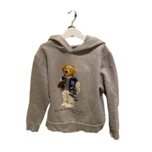 Grå hoodie Polo Bear Ralph Lauren - Snygg grå hoodie från Ralph Lauren med ikoniska Polo Bear-trycket framtill.Storlek 10-12 år. Tröjan har huva som är gjord i mjuk bomullsmix. En liten defekt på trycket men de är inget man märker på avstånd. Perfekt för en chill och sportig look med streetkänsla. Priset är ej hugget sten. Hör av er vid frågor eller funderingar! 🧸