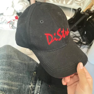 Svart keps med röd DiStra-brodyr - Svart keps med böjd skärm och röd brodyr framtill med texten 'DiStra' samt webbadress bak. Justerbar rem baktill och röd detalj längs skärmens kant. Snygg och enkel design, perfekt för dig som gillar streetwear. 89 för båda.