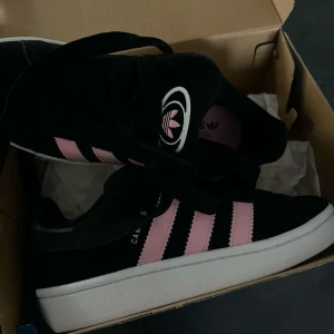 Adidas Campus svart/rosa sneakers - Snygga Adidas Campus sneakers i svart mocka med klassiska rosa ränder på sidorna och vit sula. Skorna har snörning och en ikonisk Adidas-logga på plösen. Perfekta för dig som vill sticka ut med en cool färgkombo och retrovibe.