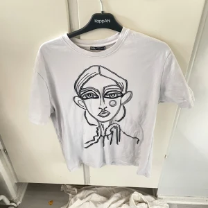 Vit t-shirt med broderad ansikte Zara - Vit t-shirt från Zara med ett broderat ansikte i svart på framsidan. Klassisk rund halsringning och korta ärmar. T-shirten är i mjuk bomull och har en clean, modern vibe som passar perfekt till jeans eller shorts.