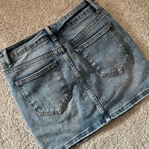 Blå jeanskjol - Chelsea - Blå jeanskjol i storlek 34 köpt från Chelsea med märket Miss Bon med klassisk femficksdesign, bälteshällor och knappstängning framtill. Kjolen är kort och har en rak passform.