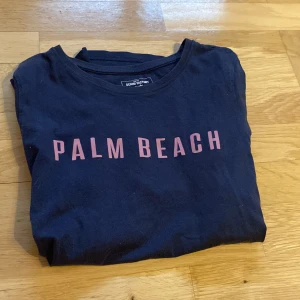 Svart t-shirt Palm Beach Denim Factory - Svart t-shirt från Denim Factory med trycket 'PALM BEACH' i rosa bokstäver på bröstet. Klassisk rund halsringning och korta ärmar. Skön och enkel stil som passar perfekt till jeans eller shorts.