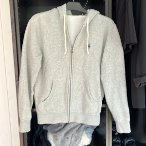 Grå hoodie från Polo Ralph Lauren - Snygg grå hoodie från Polo Ralph Lauren med dragkedja, huva och vita snören. Klassisk broderad logga i mörkblått på bröstet. Tillverkad i mjuk bomullsmix, perfekt för chill dagar eller när du vill ha en clean och enkel stil. Passar mig som är 173.