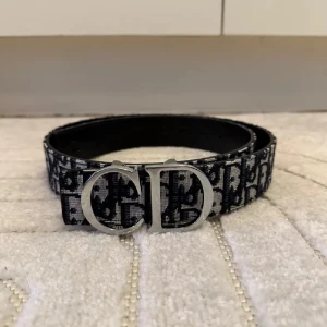 Svart och vit Dior bälte med CD-spänne - Snyggt bälte från Dior med ikoniskt CD-spänne i silver. Bältet har svart och vit monogramdesign med tydligt Dior-mönster längs hela bandet. Materialet är textil med insida i skinn, perfekt för att lyfta vilken outfit som helst.