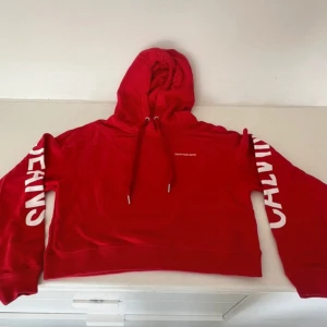 Röd hoodie från Calvin Klein Jeans - Snygg röd hoodie från Calvin Klein Jeans med vit logga på bröstet och stora tryckta bokstäver på ärmarna. Tröjan har huva med snörning och är croppad i modellen. Perfekt för dig som gillar streetstyle och vill sticka ut.