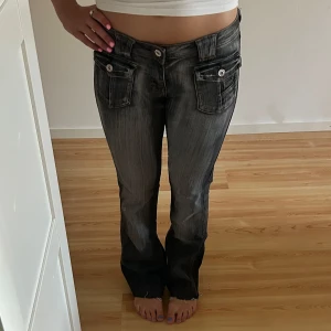 Grå jeans med stora fickor - Snygga grå jeans med slitningseffekt och stora fickor med lock och knappar både fram och bak. Byxorna har låg midja och raka ben med lätt bootcut. Perfekta för dig som gillar streetstyle och detaljer.