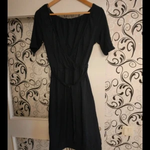 Svart omlottklänning med knytband - En svart omlottklänning med knytband i midjan och korta ärmar. Klänningen har en v-ringad omlottfront och är knälång, vilket ger en klassisk och stilren look. Perfekt för dig som gillar enkel men snygg design.