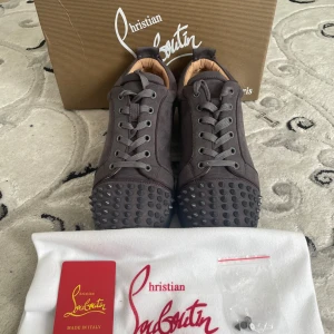 Christian Louboutin sneakers med nitar - Mörkgrå sneakers från Christian Louboutin i mocka med ikoniska röda sulor och coola nitar på tån. Skorna har snörning och en rund tå, samt lyxiga detaljer som gör dem till ett statement. Perfekta för dig som vill sticka ut med exklusiv stil.