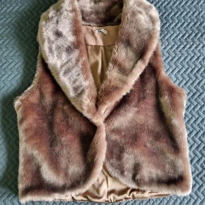 Beige pälsväst med peace-märke - Säljer en supermjuk och fluffig beige pälsväst med bred krage. Baksidan har ett stort peace-märke i mocka och nitar som ger en cool retrovibe. Utsidan är i fuskpäls med bruna och beige toner, insidan är slät och bekväm. Perfekt för dig som vill sticka ut!