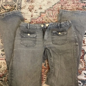 Grå jeansbyxor från Gina Tricot - Snygga grå jeansbyxor från Gina Tricot med raka ben och coola fickor framtill med lock och silverfärgade knappar. Klassisk femficksmodell och bälteshällor. Perfekta för en chill och stilren look.