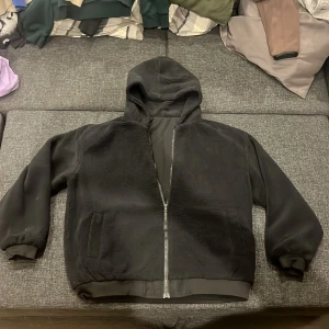 Svart teddyjacka med huva - Nypris 850kr. Mysig svart teddyjacka med huva och dragkedja framtill. Jackan har en enkel design med ribbade muddar och en ficka på sidan. Perfekt för kyliga dagar när du vill vara både bekväm och stilren.