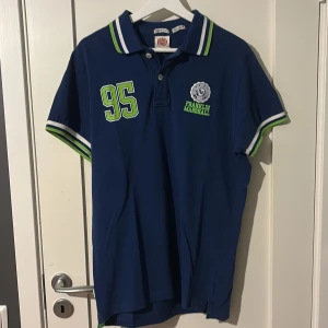 Blå pikétröja Franklin & Marshall - Snygg blå pikétröja från Franklin & Marshall med gröna och vita detaljer. Stort tryck med siffran 95 på både fram- och baksida, samt broderad logga på bröstet. Krage och ärmslut har coola ränder i grönt och vitt. Perfekt för en sportig look.