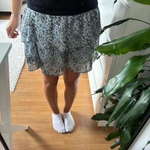 Leopardmönstrad volangkjol Chelsea - Supersöt kort kjol från Chelsea i ljusblått med svart och grått leopardmönster. Kjolen har smockad midja och dubbla volanger som ger en flowy känsla. Perfekt för dig som gillar att sticka ut med coola mönster och mjuka material.