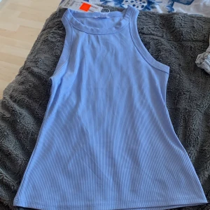 Ljusblå ribbad linnetopp - Säljer en ljusblå ribbad linnetopp med hög halsringning och ärmlös design. Perfekt för varma dagar och enkel att styla till jeans eller kjol. Toppens material är mjukt och stretchigt för en bekväm passform.