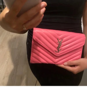 Rosa YSL kuvertväska i skinn - (Copy1 från Turkiet) Säljer en snygg rosa kuvertväska från YSL i quiltat skinn med ikoniskt YSL-emblem i silver framtill. Väskan har ett stilrent, kuvertformat lock och trendig chevronmönstring. 