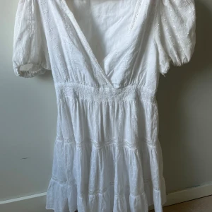 Vit kort klänning från Zara XS - Supersöt vit kort klänning från Zara med broderade detaljer, puffärmar och v-ringning. Klänningen har omlottliv och volang i kjolen, perfekt för sommarens alla tillfällen. Tillverkad i bomull för en luftig och skön känsla.