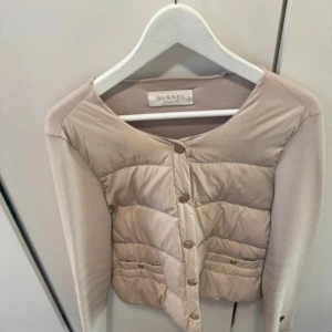 Beige dunväst från Busnel - Snygg beige dunväst från Busnel med quiltad front och knappar i guldton. Västen har en stilren, rund halsringning och är tillverkad i en mix av dun och stickat material på sidorna. Perfekt för lager-på-lager och ger en clean look.